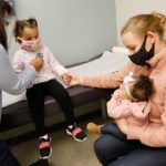 A female pediatrician checking on a little girl's heartbeat while a mother holds her little girl's hand and holds her newborn child in her other arm.