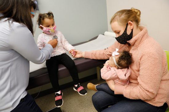 A female pediatrician checking on a little girl's heartbeat while a mother holds her little girl's hand and holds her newborn child in her other arm.