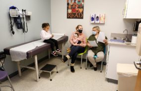 A female specialist is speaking to a mother who is holding her newborn baby while her toddler sits on the exam table in the exam room.
