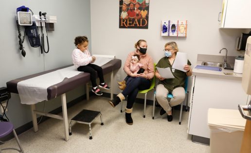 A female specialist is speaking to a mother who is holding her newborn baby while her toddler sits on the exam table in the exam room.