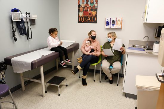 A female specialist is speaking to a mother who is holding her newborn baby while her toddler sits on the exam table in the exam room.