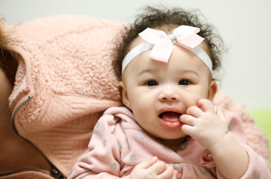 A closeup of a baby girl wearing a pink headband with a bow as she looks into the camera.