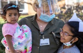 A female specialist is wearing a mask while holding a toddler in her arms in one hand while looking down and holding a young girl next to her.