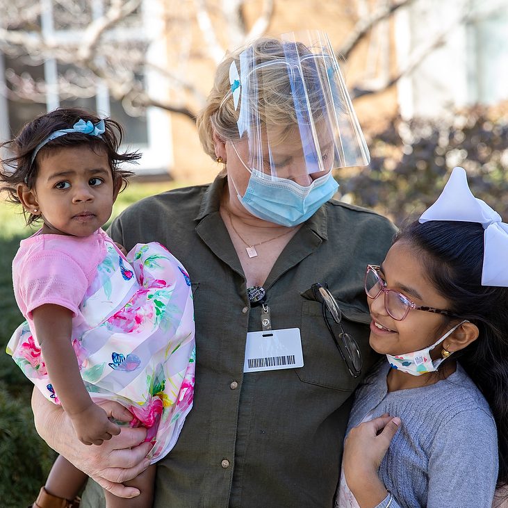 A female specialist is wearing a mask while holding a toddler in her arms in one hand while looking down and holding a young girl next to her.