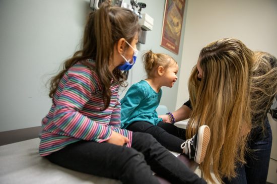 A female specialist making a toddler laugh while another toddler sits next to her wearing a mask.
