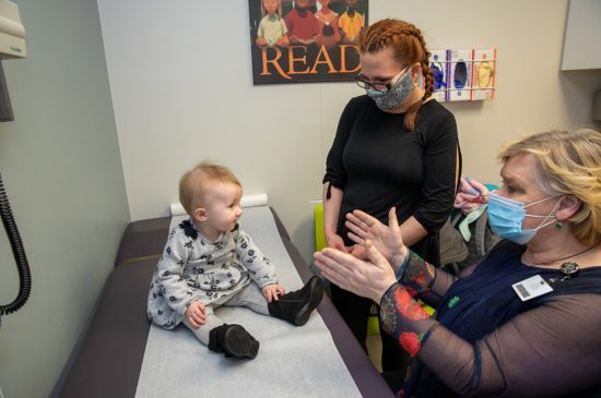 A female specialist wearing a mask is sitting down making a toddler comfortable while the mother also wearing a mask watches while standing in an exam room.