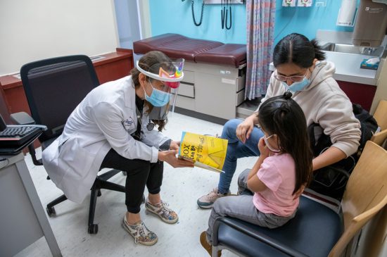 A female pediatrician speaking with a little girl and her mother in an exam room, all wearing face masks.