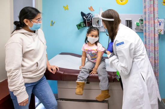 A female pediatrician is checking a toddler's heart rate while the mother is standing next to the toddler all wearing masks.