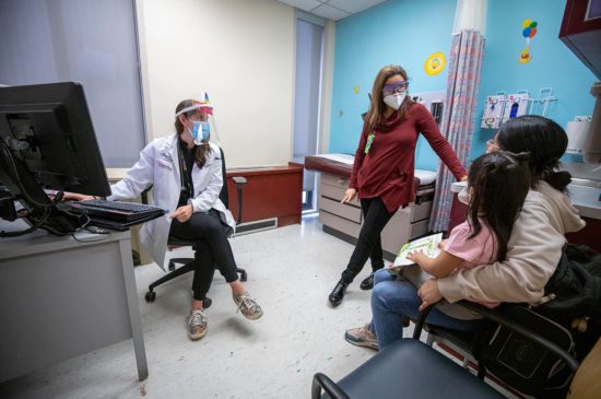 A female pediatrician sitting next to a female specialist that is standing talking with a mother while she holds her little girl in her lap.