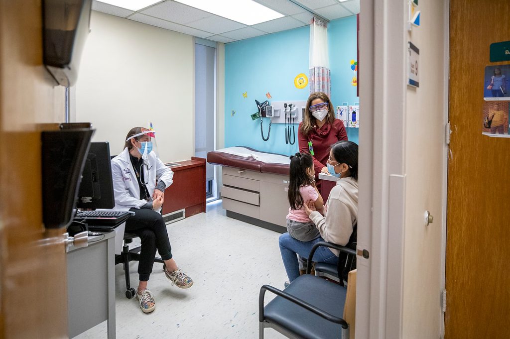 A female specialist standing and a female pediatrician sitting listening to a mother speak while her child sits on her lap in an exam room.