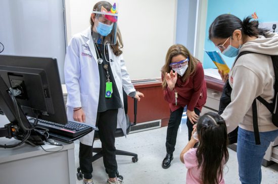 A female pediatrician standing next to a female specialist who is bent over speaking to a toddler who is holding hands with her mother.