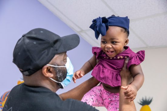 A father wearing a face mask is holding his little girl up in the air while she is smiling.