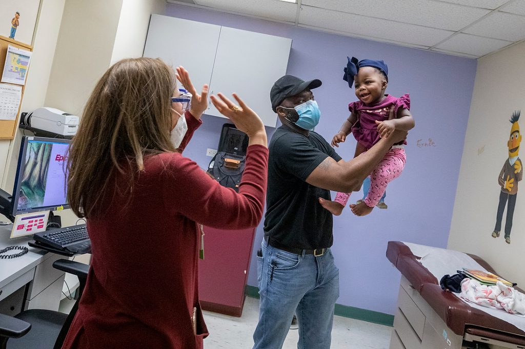 A father holding his little daughter in the air while a female specialist is holding her hands in the air, making his child laugh in an exam room.