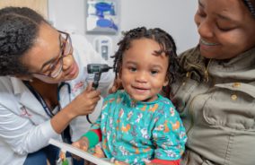 A toddler looking at the camera while sitting in his mothers lap as a female pediatrician looks into his ears.