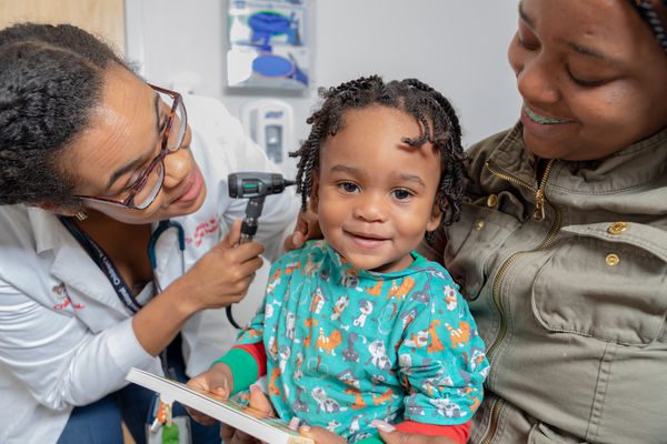 A toddler looking at the camera while sitting in his mothers lap as a female pediatrician looks into his ears.