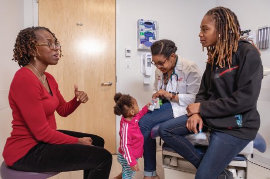 A female specialist is talking to a parent while a toddler looks at a female pediatrician’s badge all while sitting in an exam room.