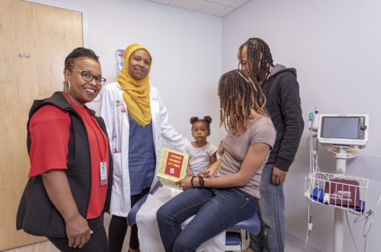 A female specialist and female pediatrician smile at the camera while a couple show their toddler a book in the exam room.