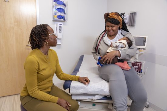 A female specialist is sitting down talking with a mother while she holds her newborn smiling in an exam room.