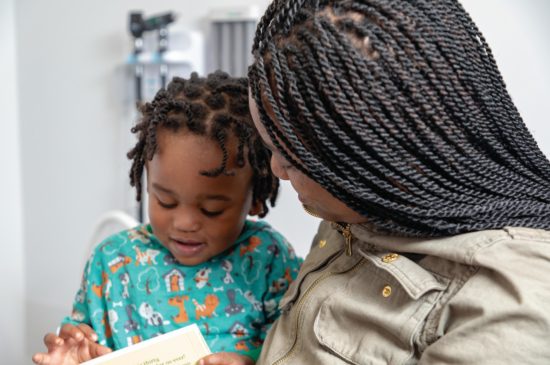 Toddler sits on mom's lap reading a book at the pediatrician's office