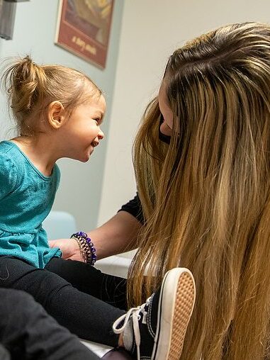 A female specialist making a toddler laugh while another toddler sits next to her wearing a mask.